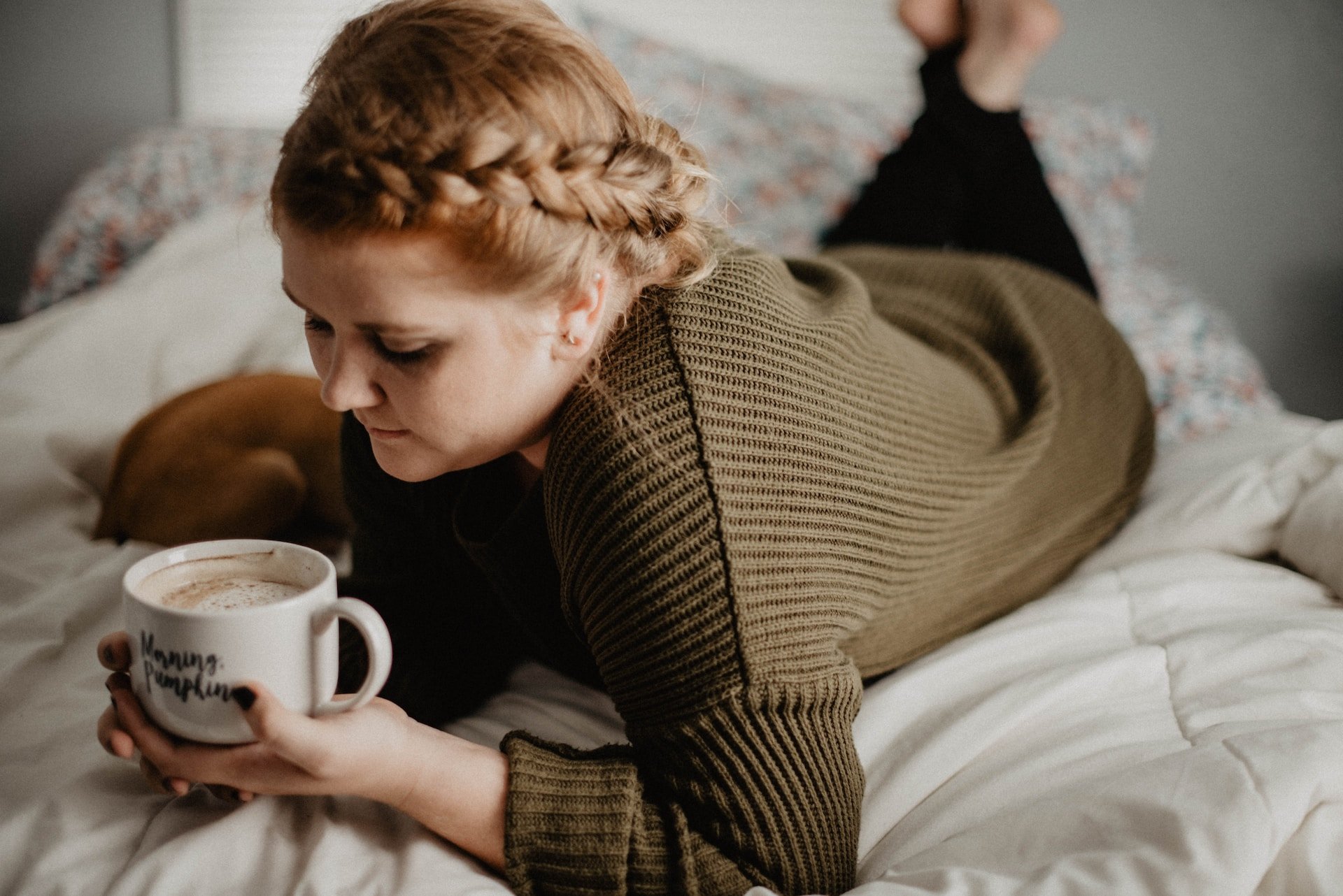 Red-haired woman with braid lounging in bed with mug of warm drink, dog snuggled up in blurred background